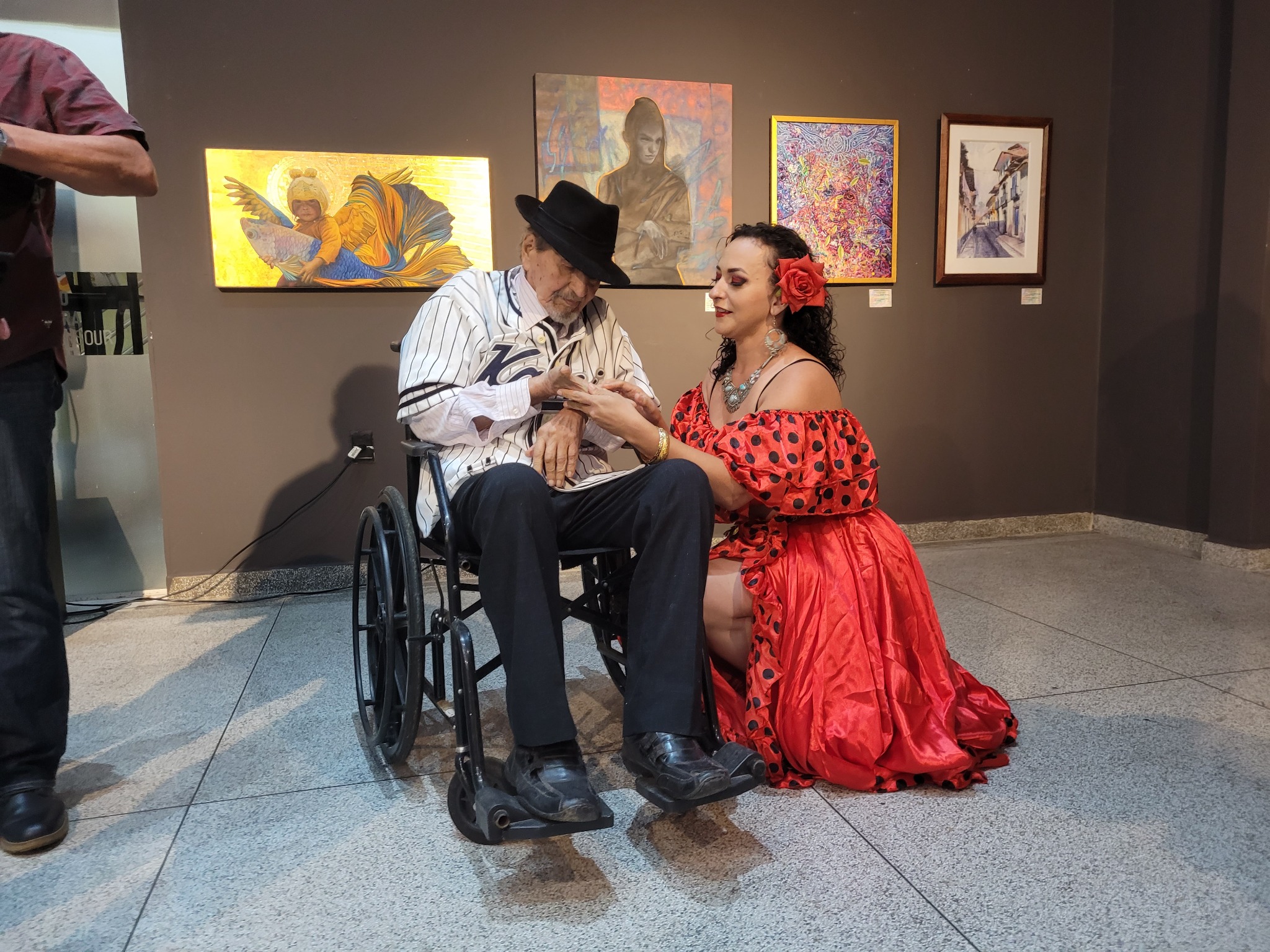 Paula Santos de vermelho olhando a mão de Rivas Plata, sentado em uma cadeira de rodas, diante de pinturas.